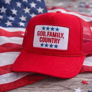May include: Red trucker hat with a white patch featuring the text "GOD, FAMILY, COUNTRY" and blue stars. The hat is set against an American flag background.