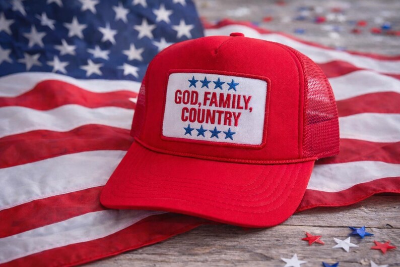 May include: Red trucker hat with a white patch featuring the text "GOD, FAMILY, COUNTRY" and blue stars. The hat is set against an American flag background.