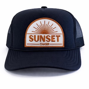 May include: Navy blue trucker hat with a curved brim and mesh back. The front features a patch with a sunset design and the words "SUNSET CHASER" in orange lettering. A stylish accessory for outdoor enthusiasts.