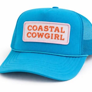 May include: A turquoise trucker hat with a mesh back and a curved brim. The hat features a rectangular patch with the words "COASTAL COWGIRL" in orange lettering on a white background with a pink border.