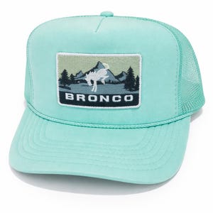 May include: A light teal trucker hat with a mesh back and a curved brim. The front features a rectangular patch with a mountain and tree design, and the word "BRONCO" in white letters.