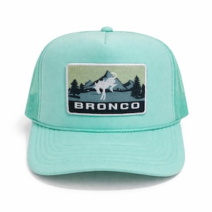 May include: A light teal trucker hat with a mesh back and a curved brim. The front features a rectangular patch with a mountain scene, a white animal silhouette, and the word "BRONCO" in bold white letters.