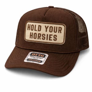 May include: A brown trucker cap with a mesh back and a curved peak. The cap has a rectangular patch with the words "HOLD YOUR HORSIES". A small brand tag is visible. The cap is a popular style.