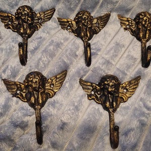 May include: A set of five identical wall hooks made of dark metal with a distressed gold finish. Each hook features a cherub figure with outstretched wings and a single curved hook extending from the bottom, designed for mounting with two screw holes.