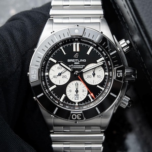 May include: A stainless steel Breitling Chronomat watch with a black dial and white sub-dials. The watch features a tachymeter bezel and a date display at the 6 o'clock position. The watch is on a stainless steel bracelet.