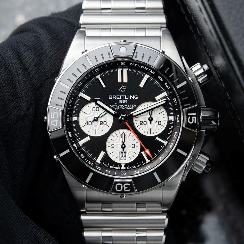 May include: A stainless steel Breitling Chronomat watch with a black dial and white sub-dials. The watch features a tachymeter bezel and a date display at the 6 o'clock position. The watch is on a stainless steel bracelet.