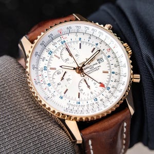 May include: A gold-toned Breitling Chronometre Navitimer watch with a white face and brown leather strap. The watch features multiple subdials, a date display, and a rotating bezel. The watch face displays the text "BREITLING 1884 CHRONOMETRE NAVITIMER".