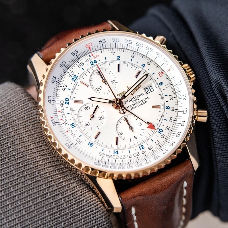May include: A gold-toned Breitling Chronometre Navitimer watch with a white face and brown leather strap. The watch features multiple subdials, a date display, and a rotating bezel. The watch face displays the text "BREITLING 1884 CHRONOMETRE NAVITIMER".