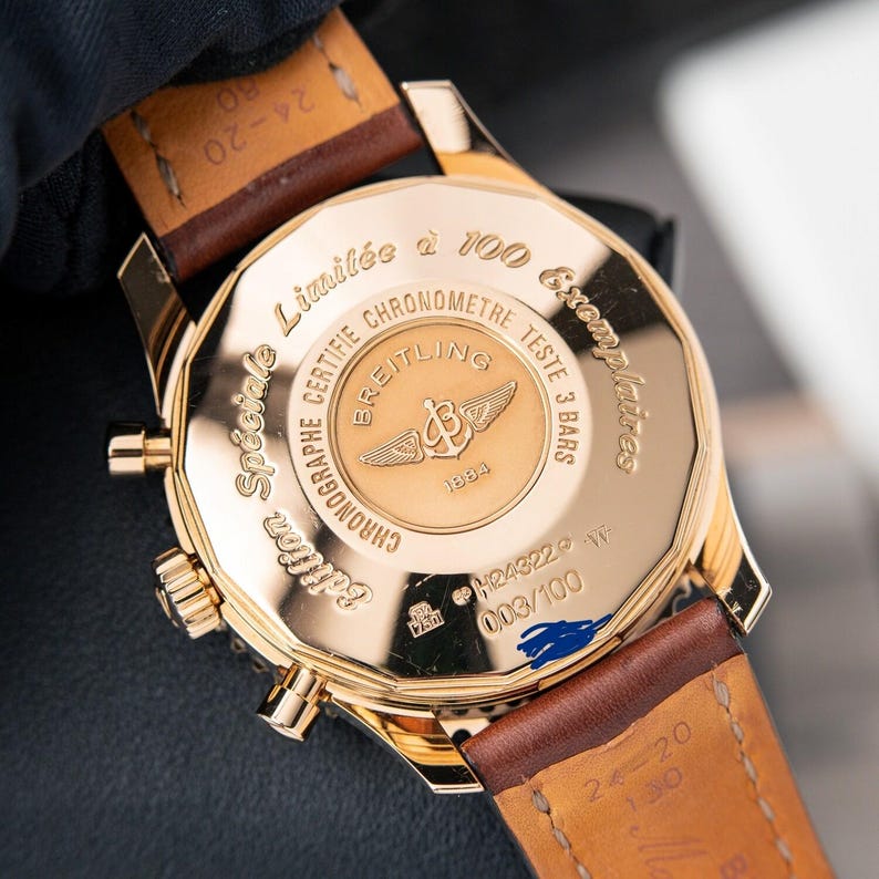 May include: A gold-toned Breitling chronograph watch with a brown leather strap. The watch back features engraved text including "Limited to 100 Exemplaires" and "Chronographe Certifie". The watch is a special edition, with the number 003/100 visible.