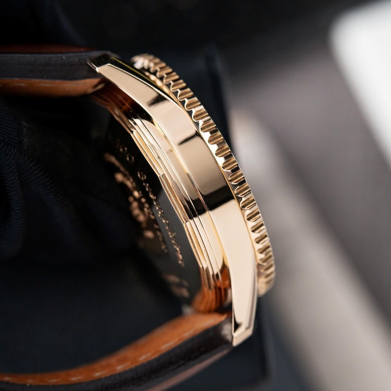 May include: Close-up of a gold-toned wristwatch. The watch features a textured bezel and a brown leather strap. The watch face has engraved details. The watch is set against a dark background.