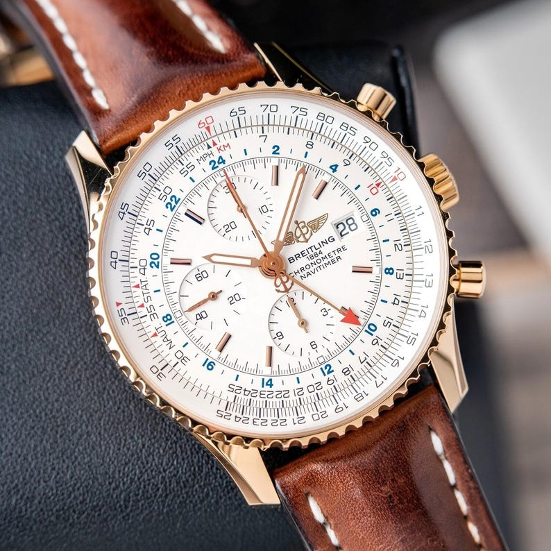 May include: A gold-toned Breitling Navitimer chronograph watch with a white face, featuring multiple sub-dials and a date display. The watch has a brown leather strap with white stitching. The watch face displays the text "BREITLING 1884 CHRONOMETRE NAVITIMER".