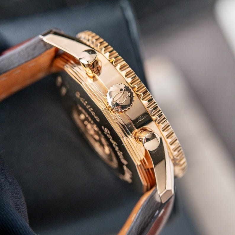 May include: Close-up of a gold-toned wristwatch with a brown leather band. The watch features a detailed bezel and multiple dials. The watch face has the brand logo and text. The watch band is approximately 0.75 inches wide.