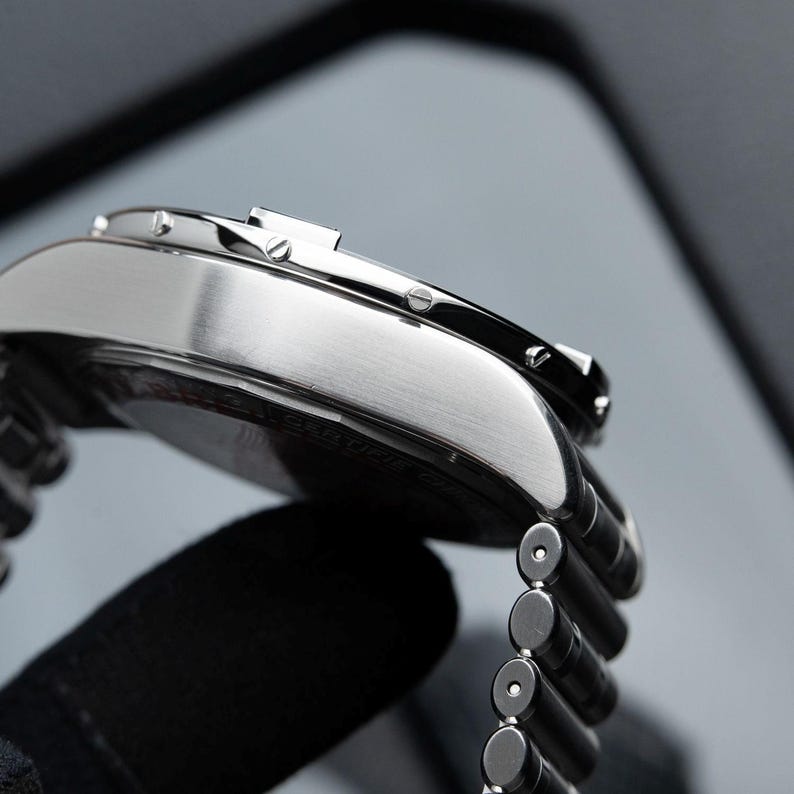 May include: Close-up of a silver wristwatch with a brushed metal finish. The watch has a black dial and a stainless steel band. The watch is held by a gloved hand.