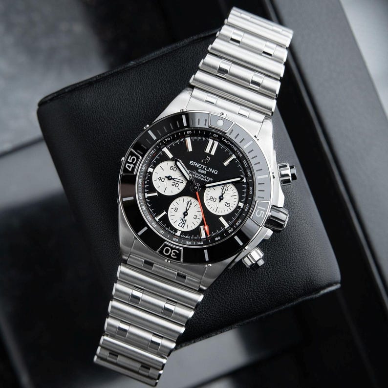 May include: A Breitling chronograph watch with a black dial, white sub-dials, and a stainless steel band. The watch features a rotating bezel with minute markers and a date display. The watch is displayed on a black surface.
