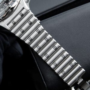May include: Close-up of a silver-toned watch band. The band is made of rectangular metal links with square cutouts. The watch band is resting on a black surface. The watch face is visible in the upper left corner.