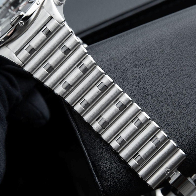 May include: Close-up of a silver-toned watch band. The band is made of rectangular metal links with square cutouts. The watch band is resting on a black surface. The watch face is visible in the upper left corner.