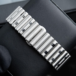 May include: A silver-toned Breitling watch band with a series of rectangular links. The band is made of metal and features the Breitling logo and brand name. The band is resting on a black leather surface.