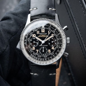 Könnte beinhalten: Eine Breitling Navitimer Uhr mit schwarzem Zifferblatt, silbernem Gehäuse und schwarzem Lederarmband. Das Zifferblatt verfügt über mehrere Hilfszifferblätter, Ziffern und das Breitling-Logo. Die Uhr hat eine drehbare Lünette.