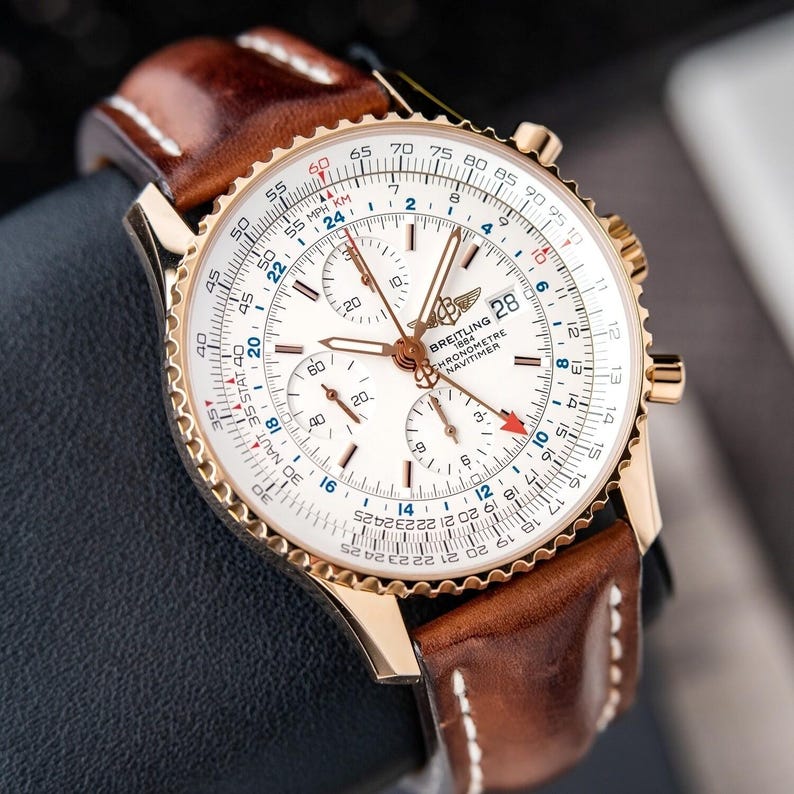 May include: A gold-toned Breitling Navitimer watch with a white face, featuring multiple subdials and a date display. The watch has a brown leather strap with white stitching. The bezel has a detailed scale, and the watch is a classic timepiece.