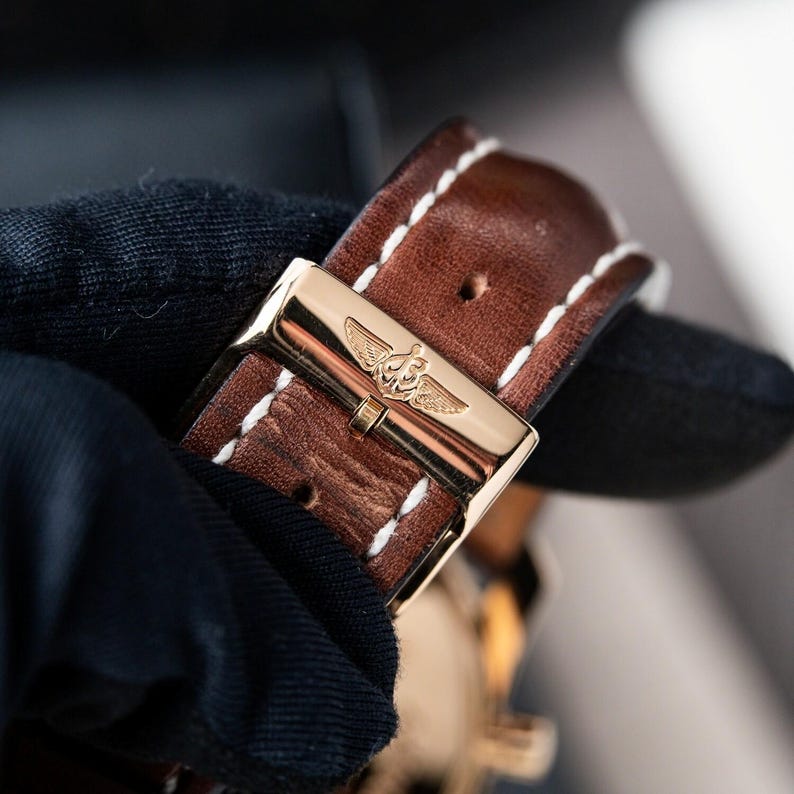 May include: Close-up of a brown leather watch strap with white stitching. The strap features a gold-colored buckle with a detailed emblem. The watch strap is a rich brown color.