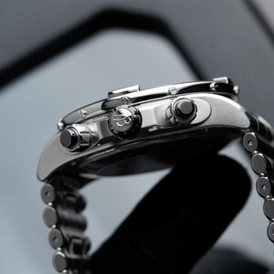 May include: Close-up of a silver wristwatch with a stainless steel band. The watch features a black dial, black buttons, and a detailed bezel. The watch is a luxury item, showcasing fine craftsmanship and design.