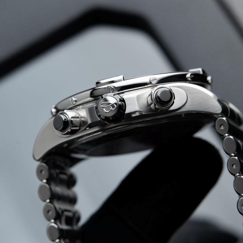 May include: Close-up of a silver wristwatch with a stainless steel band. The watch features a black dial, black buttons, and a detailed bezel. The watch is a luxury item, showcasing fine craftsmanship and design.