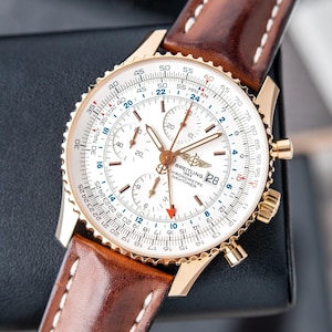 May include: A gold-toned Breitling Navitimer watch with a white face and brown leather strap. The watch features multiple subdials, a date window, and a rotating bezel. The strap is brown leather with white stitching.