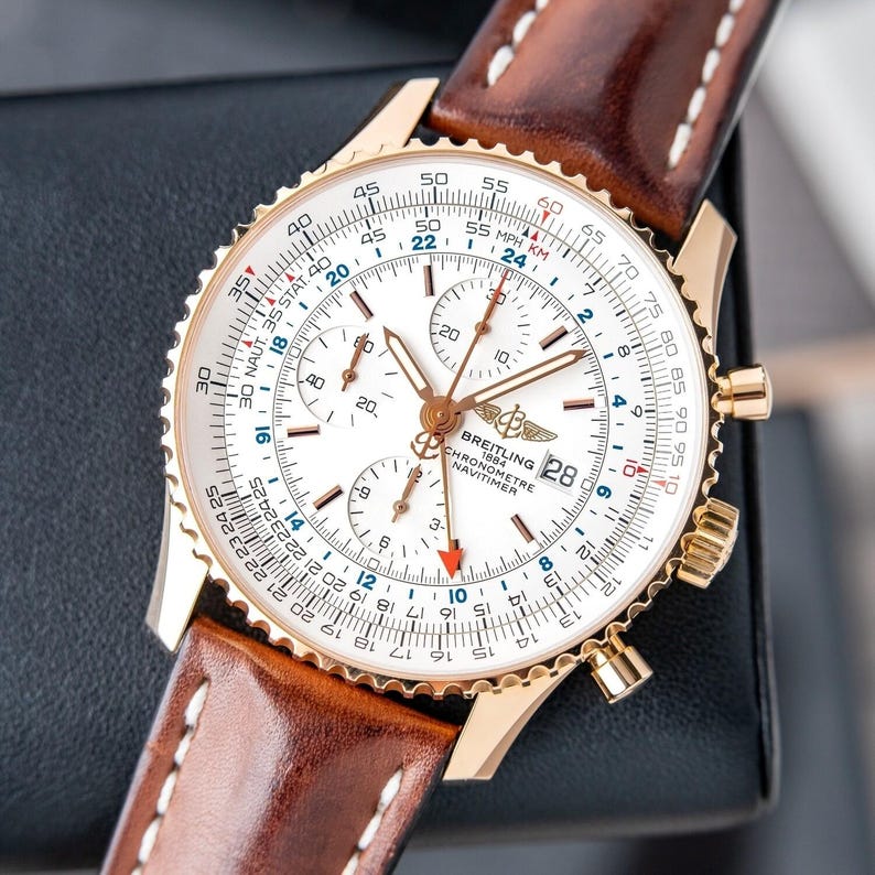 May include: A gold-toned Breitling Navitimer watch with a white face and brown leather strap. The watch features multiple subdials, a date window, and a rotating bezel. The strap is brown leather with white stitching.