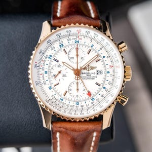 May include: A gold-toned Breitling Navitimer watch with a white face, featuring multiple subdials and a date display. The watch has a brown leather strap with white stitching. The watch face displays various scales and markings.