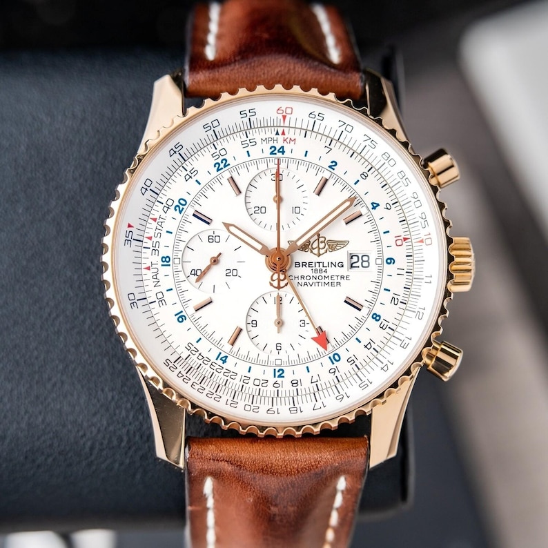 May include: A gold-toned Breitling Navitimer watch with a white face, featuring multiple subdials and a date display. The watch has a brown leather strap with white stitching. The watch face displays various scales and markings.