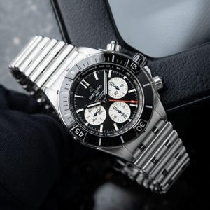 May include: A stainless steel Breitling chronograph watch with a black dial and three sub-dials. The watch features a tachymeter bezel and a silver-tone bracelet. The watch is displayed on a black surface.