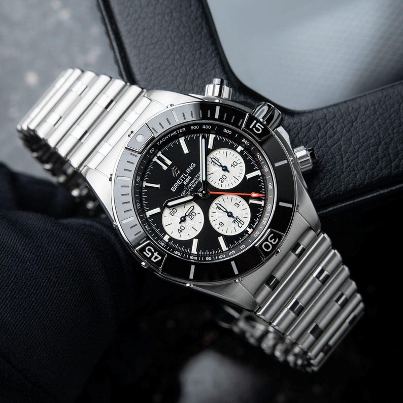 May include: A stainless steel Breitling chronograph watch with a black dial and three sub-dials. The watch features a tachymeter bezel and a silver-tone bracelet. The watch is displayed on a black surface.