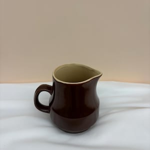 May include: A small, dark brown ceramic pitcher with a curved spout and a rounded handle. The interior is a light beige color. The pitcher is set against a white background.