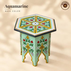 May include: A hexagonal side table with an aquamarine base color, featuring hand-painted floral motifs in red, green, and yellow. The top surface displays a geometric pattern with a central white flower, while the legs have arched cutouts.