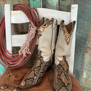 May include: A pair of cream-colored cowboy boots with snake print detailing on the foot and upper shaft. The boots are set on a brown leather surface with fringe and decorative metal accents. A red rope and white chair are in the background.