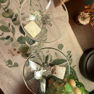 May include: Two clear glass coupes with wide, circular bowls and faceted stems. Each glass features a smooth, transparent rim and a distinct geometric base design. A small, square, off-white adhesive label is attached to the interior of each bowl.