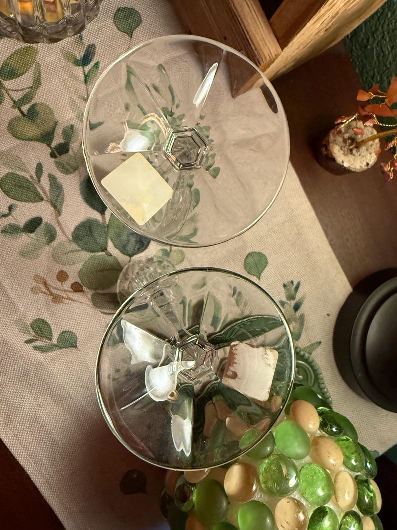 May include: Two clear glass coupes with wide, circular bowls and faceted stems. Each glass features a smooth, transparent rim and a distinct geometric base design. A small, square, off-white adhesive label is attached to the interior of each bowl.