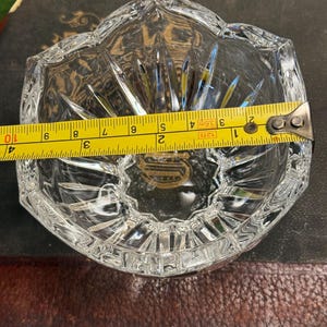 May include: A clear, faceted crystal ashtray with a starburst design. The ashtray is approximately 4 inches wide. The edges are cut with a decorative pattern. The ashtray is sitting on a dark surface.