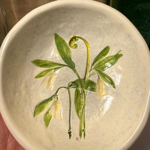 May include: A small, off-white ceramic bowl with a hand-painted floral design. The design features green leaves and yellow-white bell-shaped flowers. The bowl is round and has a slightly textured surface. The bowl is held in a hand.