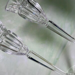 May include: A pair of clear glass candle holders featuring a fluted, bell-shaped top section with a faceted rim. Each holder has a slender, multi-sided stem that transitions into a smooth, circular base, providing a stable foundation for candles.