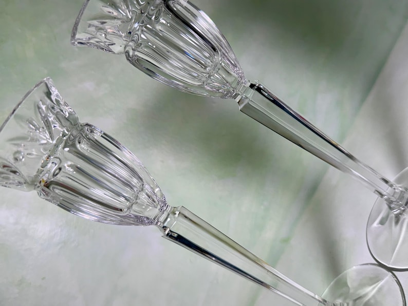 May include: A pair of clear glass candle holders featuring a fluted, bell-shaped top section with a faceted rim. Each holder has a slender, multi-sided stem that transitions into a smooth, circular base, providing a stable foundation for candles.