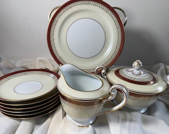 May include: A cream-colored porcelain tea set featuring a deep red border with a gold leaf pattern. The set includes a large serving plate, a stack of small saucers, a creamer pitcher with a handle, and a lidded sugar bowl with two side handles.