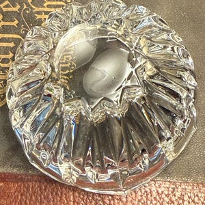 May include: A clear, faceted glass ashtray with a scalloped edge. The ashtray has a circular shape with a deep center and a textured, reflective surface. The ashtray is sitting on a dark surface.