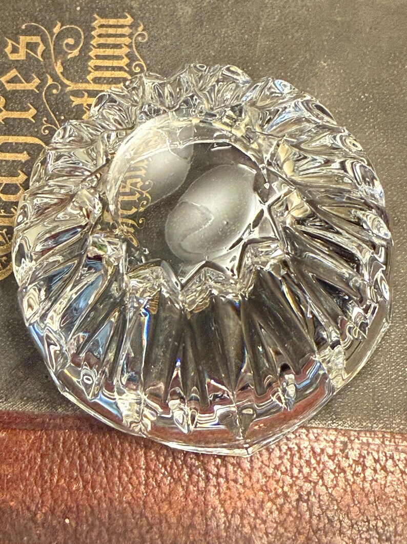 May include: A clear, faceted glass ashtray with a scalloped edge. The ashtray has a circular shape with a deep center and a textured, reflective surface. The ashtray is sitting on a dark surface.