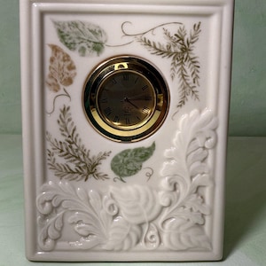 May include: A rectangular white ceramic mantel clock featuring a central circular gold-toned clock face with Roman numerals. The surface is decorated with embossed leaf patterns at the bottom and painted green and brown leaf motifs above.