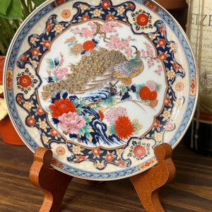 Puede incluir: Un plato decorativo de porcelana con un diseño de pavo real. El plato tiene un centro blanco con una ilustración detallada de un pavo real posado en una rama, rodeado de flores y follaje coloridos. El borde está adornado con un patrón floral azul y dorado.