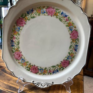 Könnte beinhalten: Cremefarbener Teller mit silberner Umrandung und gewelltem Rand. Der Teller zeigt ein Kreuzstich-Blumenmuster in Rosa, Blau, Gelb und Grün um die Mitte. Der Teller wird auf einem transparenten Kunststoffständer präsentiert.