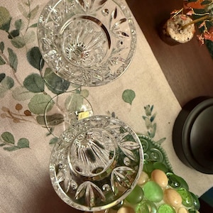 May include: Two clear crystal candle holders featuring a faceted, starburst-like pattern around the rim and vertical fluting along the interior bowl. Each piece has a circular base and a small, rectangular gold-colored sticker on the stem.