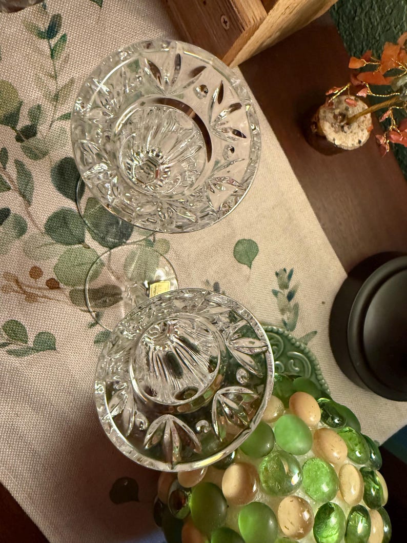May include: Two clear crystal candle holders featuring a faceted, starburst-like pattern around the rim and vertical fluting along the interior bowl. Each piece has a circular base and a small, rectangular gold-colored sticker on the stem.