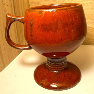 May include: A handmade, ceramic mug with a unique goblet-style base. The mug features a rich, glossy, reddish-brown glaze with darker accents. It has a curved handle and a wide, open top, perfect for coffee or tea.
